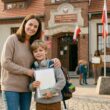 Uśmiechnięta matka obejmuje syna z plecakiem i słuchawkami przed wejściem do zabytkowego budynku polskiej szkoły podstawowej, ozdobionego flagami narodowymi. W tle widać inne dzieci i nauczycielkę na dziedzińcu.